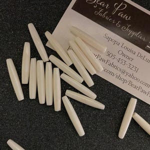 May include: A collection of off-white, smooth, cylindrical beads. The beads are arranged in a pile on a black surface. The beads are approximately 1 inch in length.