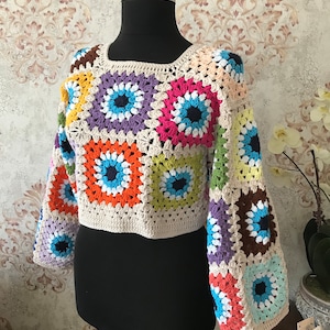 Puede incluir: Top de ganchillo con diseño de cuadrados multicolores. El top presenta un cuello cuadrado y mangas largas. Destacan colores vibrantes como el azul, naranja y rosa, sobre un fondo crema. La prenda se exhibe en un maniquí negro.