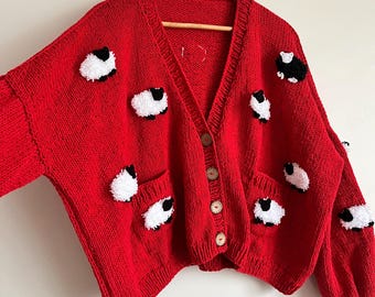 Könnte beinhalten: Eine leuchtend rote Strickjacke mit V-Ausschnitt, zwei Taschen und Holzknöpfen. Der Pullover ist mit flauschigen, weißen und schwarzen Schafapplikationen verziert. Das Kleidungsstück hängt an einem Holzkleiderbügel.