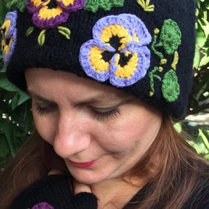 May include: Black knitted hat with a pom pom on top and embroidered purple, yellow, and blue pansies. The hat is worn by a person with long brown hair.