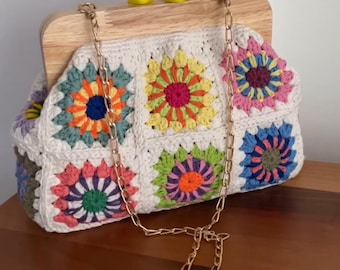 May include: A handmade crochet bag with a wooden frame and gold chain strap. The bag features colorful floral squares in shades of orange, yellow, pink, blue, and green against a white background. Two yellow cube-shaped accents sit on top of the wooden frame.