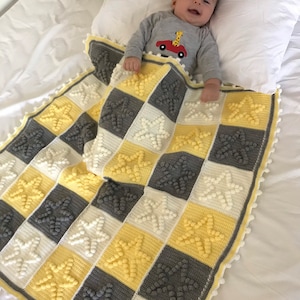 May include: A baby is lying on a white bed with a yellow, white and grey crocheted blanket with a star pattern. The blanket has a white pom-pom trim.