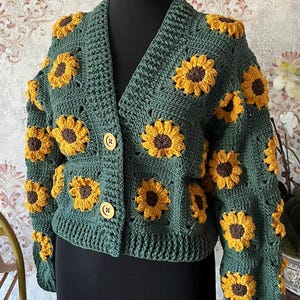 May include: A green crocheted cardigan with a V-neck and button closure. The cardigan is decorated with yellow and brown crocheted sunflowers.