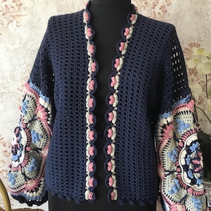 Peut inclure: Un cardigan en crochet bleu marine avec un motif ajouré délicat. Les manches présentent des motifs floraux complexes dans des tons de rose, bleu et blanc.