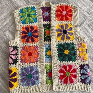 May include: A sleeveless waistcoat constructed from multiple crocheted granny squares, each featuring a vibrant multi-coloured flower motif. The garment is bordered with a cream-coloured yarn trim and showcases a variety of colours including red, orange, yellow, green, blue, purple, and pink.