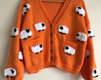 Könnte beinhalten: Orangefarbener Strickcardigan mit weißen, flauschigen Schaf-Applikationen. Der Cardigan hat einen V-Ausschnitt und einen Knopfverschluss.