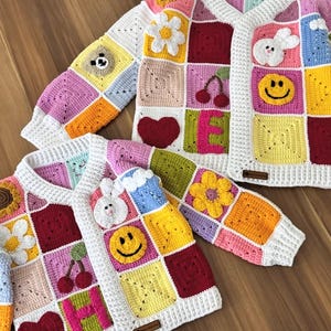 May include: Three colorful, crocheted cardigans with a patchwork design. Each cardigan features a variety of squares in different colors, with appliqued flowers, smiley faces, cherries, and hearts. The cardigans have white trim and are displayed on a wooden surface.