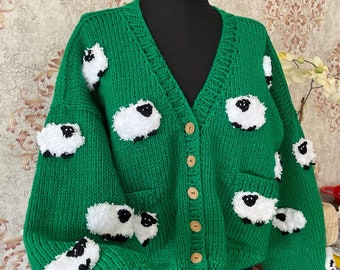 Könnte beinhalten: Grüne Strickjacke mit V-Ausschnitt und Knopfleiste. Die Jacke ist mit weißen, flauschigen Schaf-Applikationen verziert.