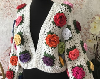 Puede incluir: Una chaqueta blanca de ganchillo con flores de ganchillo de colores. La chaqueta tiene cuello en V y mangas largas.