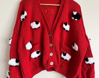 Könnte beinhalten: Rote Strickjacke mit V-Ausschnitt und Holzknöpfen. Der Pullover ist mit weißen und schwarzen Schafapplikationen verziert. Die Strickjacke hat zwei Vordertaschen und hängt an einem Holzkleiderbügel.