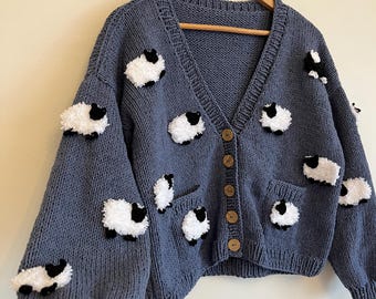 Könnte beinhalten: Eine blaue Strickjacke mit V-Ausschnitt und Holzknöpfen. Der Pullover hat mehrere Applikationen von weißen und schwarzen Schafen. Die Strickjacke hat zwei Taschen und wird auf einem Holzkleiderbügel präsentiert.