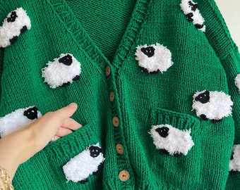 Könnte beinhalten: Ein grüner Strickcardigan mit V-Ausschnitt und Knopfleiste. Der Cardigan ist mit weißen, flauschigen Schaf-Applikationen verziert. Der Cardigan hat eine Tasche auf der linken Seite.