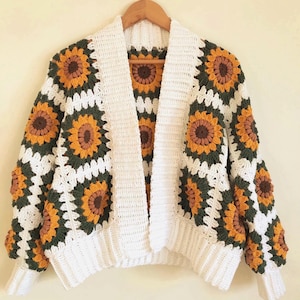 Op de afbeelding: Een handgemaakt gehaakt vest met een zonnebloemmotief. Het vest met open voorkant heeft een witte basis met vierkante zonnebloemmotieven in geel, bruin en groen. De mouwen en kraag zijn wit. Het vest wordt op een houten hanger getoond.