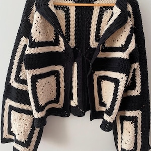 May include: A long-sleeved cardigan constructed from crocheted granny squares in a high-contrast black and cream color scheme. The garment features a repeating geometric pattern with circular floral motifs centered within each square block.