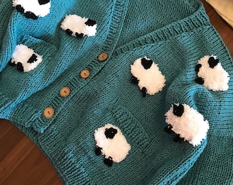 Könnte beinhalten: Ein tealblauer Strickcardigan mit V-Ausschnitt und Knopfleiste. Der Cardigan ist mit weißen und schwarzen Schaf-Applikationen verziert.