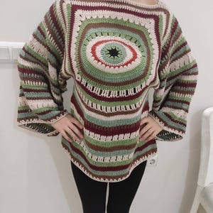 Puede incluir: Un suéter de crochet hecho a mano con un diseño de canesú cuadrado. El suéter presenta un patrón circular central con tonos de verde, marrón y rojo. Las mangas están a rayas con colores similares. El suéter es de corte holgado.
