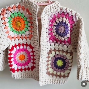 Può includere: Un cardigan all'uncinetto color crema con motivi quadrati colorati. I quadrati presentano tonalità vivaci di rosa, arancione, verde e viola. La giacca è esposta su un appendiabiti rosa.
