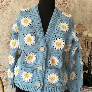 May include: A light blue crochet cardigan featuring daisy flower motifs. The cardigan has a V-neck, two pockets, and wooden buttons. The daisy flowers have white petals and yellow centers. The cardigan is displayed on a black mannequin.