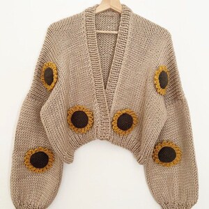 Könnte beinhalten: Ein beigefarbener Strickcardigan mit V-Ausschnitt und langen Ärmeln. Der Cardigan ist mit gehäkelten Sonnenblumenapplikationen verziert.