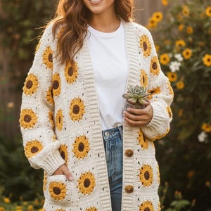 May include: Cream-colored, long crochet cardigan featuring a sunflower pattern. The cardigan has a button-up front, pockets, and a relaxed fit. The sunflowers are yellow and brown. The person is wearing a white t-shirt and blue jeans.