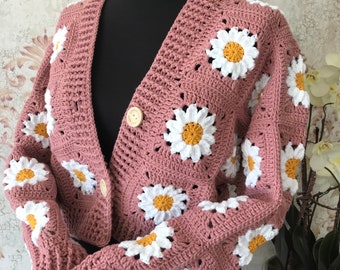 Puede incluir: Una chaqueta de punto de color rosa hecha a crochet con flores de margarita blancas y amarillas. La chaqueta tiene cuello en V y cierre de botón.