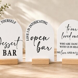 May include: Three white arched signs with black text and light wood bases. The first sign reads "TREAT YOURSELF Dessert BAR". The second sign reads "LOVE IS INTOXICATING open bar". The third sign reads "to our loved ones WHO ARE DANCING WITH US IN HEAVEN WE love AND miss YOU".