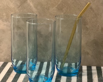 May include: Three clear glass tumblers with a light blue tint. The tumblers are sitting on a black and white striped surface. A yellow glass straw is in one of the tumblers.