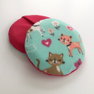 May include: Two round fleece pads. One is solid red, the other is teal with a pattern of cartoon cats, hearts, flowers, and a butterfly. The red pad is partially visible behind the patterned pad. The pads are likely for a baby or child.