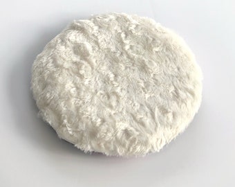May include: A round, off-white, plush cushion with a soft, textured surface. The cushion is circular and appears to be made of a faux fur material. The item is designed for comfort and decorative purposes.