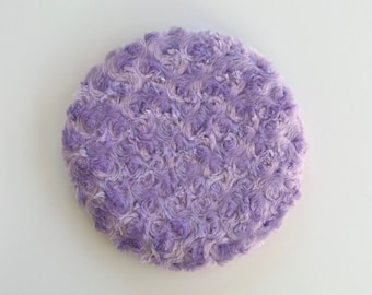 May include: A circular cushion featuring a textured, plush fabric in varying shades of light and medium purple. The surface is covered in a dense, swirling rose-like pattern created by the pile of the soft, fuzzy material, forming a uniform round shape.
