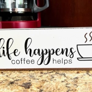 May include: White wooden sign with black text that reads "life happens coffee helps" and a black silhouette of a coffee cup with steam rising from it.