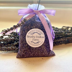 May include: A clear bag filled with dried lavender, tied with a lavender ribbon. A white sticker with the text "Brushy Hollow Farm Lavender" is on the front of the bag. Dried lavender stems are in the background.