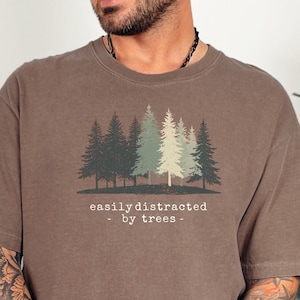 May include: Brown t-shirt with a forest graphic and the text "easily distracted by trees." The graphic features several evergreen trees in shades of green and brown. The shirt is a relaxed fit.