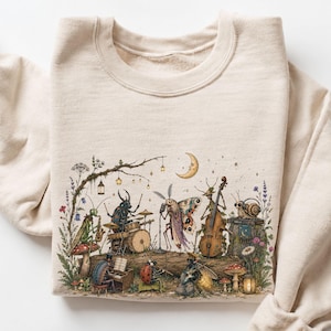 Könnte beinhalten: Ein sandfarbenes Sweatshirt mit Rundhalsausschnitt und einer detaillierten grafischen Illustration verschiedener Insekten, die in einer Waldumgebung Musikinstrumente spielen, darunter ein singender Nachtfalter, ein Käfer am Schlagzeug und eine Schnecke.