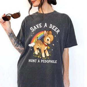 May include: Dark grey t-shirt with a cartoon deer, rainbow, stars, and the text "SAVE A DEER HUNT A PEDOPHILE" in white. The deer has a red collar and a bell. The shirt is a casual, unisex style.