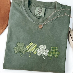 Könnte beinhalten: Grünes T-Shirt mit Rundhalsausschnitt und kurzen Ärmeln. Das Shirt zeigt vier Kleeblatt-Designs in verschiedenen Mustern und Grüntönen. Eine goldene Halskette mit Anhänger liegt auf dem Shirt. Ein brauner Hut ist sichtbar.