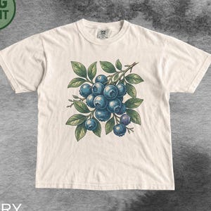 Op de afbeelding: Een ivoorkleurig t-shirt met korte mouwen en een centrale grafische illustratie van een tros donkerblauwe bosbessen aan een groene tak met bladeren. Het ontwerp is gedrukt op de voorzijde van het t-shirt met een ronde halslijn.
