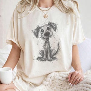 Könnte beinhalten: Cremefarbenes T-Shirt mit einer skizzierten Illustration eines Hundes. Der Hund ist sitzend dargestellt, mit großen Augen und einem verspielten Ausdruck. Das Shirt hat kurze Ärmel und eine lockere Passform, geeignet für Freizeitkleidung.