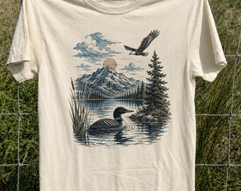 Maglietta vintage con grafica di uccelli e paesaggio, stile anni '90, colori comodi e ispirata alla natura.