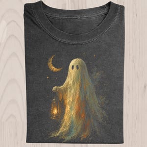 May include: A charcoal gray t-shirt featuring a painterly illustration of a pale, translucent ghost holding a glowing lantern. The ghost is positioned beneath a golden crescent moon and small stars, with soft orange and blue highlights.