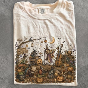 Op de afbeelding: Een crèmekleurig t-shirt met een gedetailleerde afbeelding van antropomorfe insecten die muziekinstrumenten bespelen in een bos. De illustratie bevat een bord met Happy Halloween en een ketel met de tekst Trick or Treat.