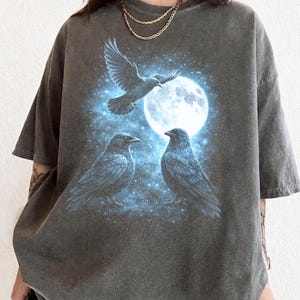 May include: Dark gray t-shirt featuring a graphic of three crows, a full moon, and a starry night sky. The crows are depicted in shades of blue and black, with one in flight. The design is centered on the shirt.