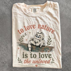 Może przedstawiać: Kremowy t-shirt z grafiką puszystego pająka z rodziny skakunowatych siedzącego na gałęzi. Napis głosi "to love nature is to love the unloved", a całość uzupełniają małe ilustracje liści i gwiazd w stonowanych odcieniach czerwieni i zieleni.