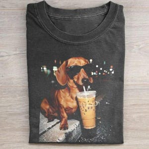 Op de afbeelding: Donkergrijze t-shirt met een bruine teckel met een zonnebril en drinkt ijskoffie. De hond zit op een stenen oppervlak met een vage achtergrond van stadslichten. De t-shirt heeft een ronde hals.