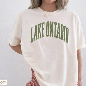 May include: An ivory short-sleeved t-shirt featuring the text LAKE ONTARIO printed in a distressed, arched green font across the chest. The garment has a crew neckline and a relaxed fit, showcasing a simple and clean design for casual wear.