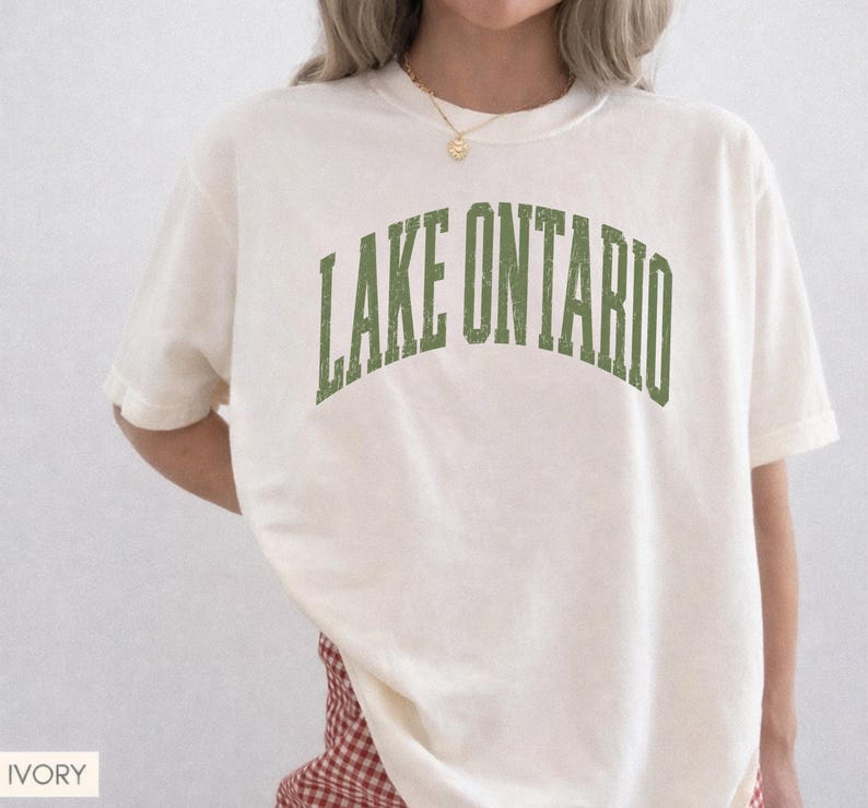 May include: An ivory short-sleeved t-shirt featuring the text LAKE ONTARIO printed in a distressed, arched green font across the chest. The garment has a crew neckline and a relaxed fit, showcasing a simple and clean design for casual wear.