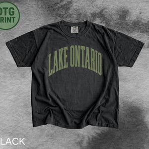 May include: A black short-sleeve crew neck t-shirt featuring the text LAKE ONTARIO in an arched, olive green collegiate-style font across the chest. The garment has a standard fit with a ribbed neckline and a small interior brand label.