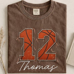Puede incluir: Una camiseta marrón de manga corta con el número 12 en un estampado de textura de balón de baloncesto naranja y bordes deshilachados. Debajo del número, el nombre Thomas está impreso en letra cursiva blanca. Tiene cuello redondo.