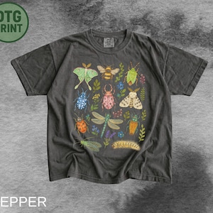 Może przedstawiać: Ciemnoszary t-shirt z krótkim rękawem z kolorowym nadrukiem graficznym przedstawiającym różne owady, w tym zieloną ćmę, trzmiela, ważkę i gąsienicę, przeplatane drobnymi ilustracjami botanicznymi kwiatów i liści.