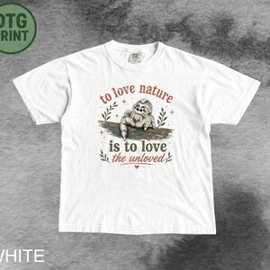 Może przedstawiać: Biały t-shirt z kr&oacute;tkim rękawem z grafiką skaczącego pająka na gałęzi. Napis "to love nature is to love the unloved" wykonany jest czerwoną i ciemnozieloną czcionką, ozdobiony małymi ilustracjami liści oraz małym czerwonym sercem.